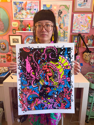 Kristen Liu-Wong holds up a copy of The Inky Depths, a vibrant, psychedelic art print featuring a pink squid beside a scientist in a colorful submersible, surrounded by marine life.